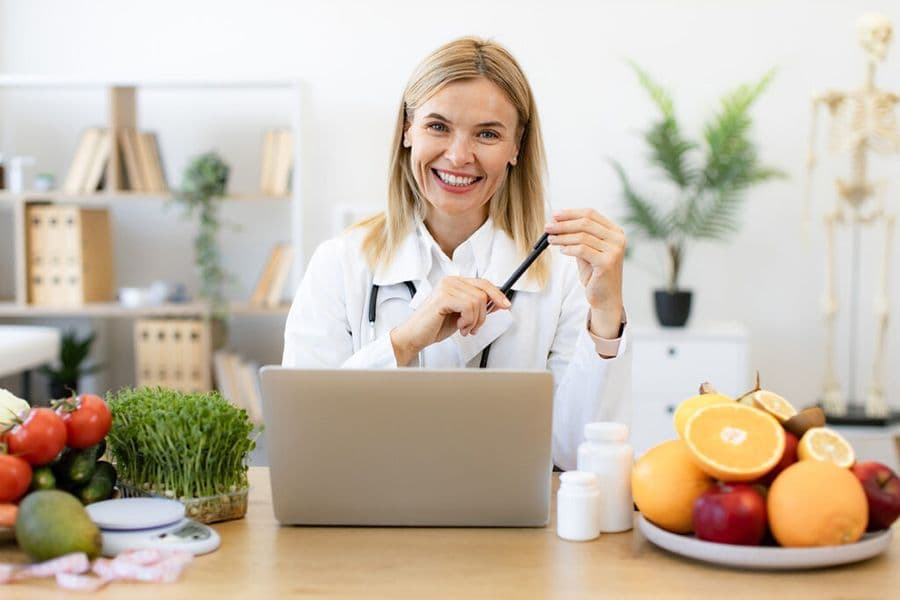 A woman in a white coat is holding a pen while standing in front of a laptop.