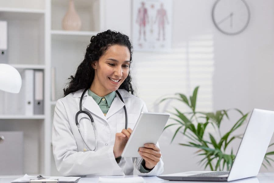 a smiling healthcare professional in a white coat using a digital tablet in a bright, modern office setting.