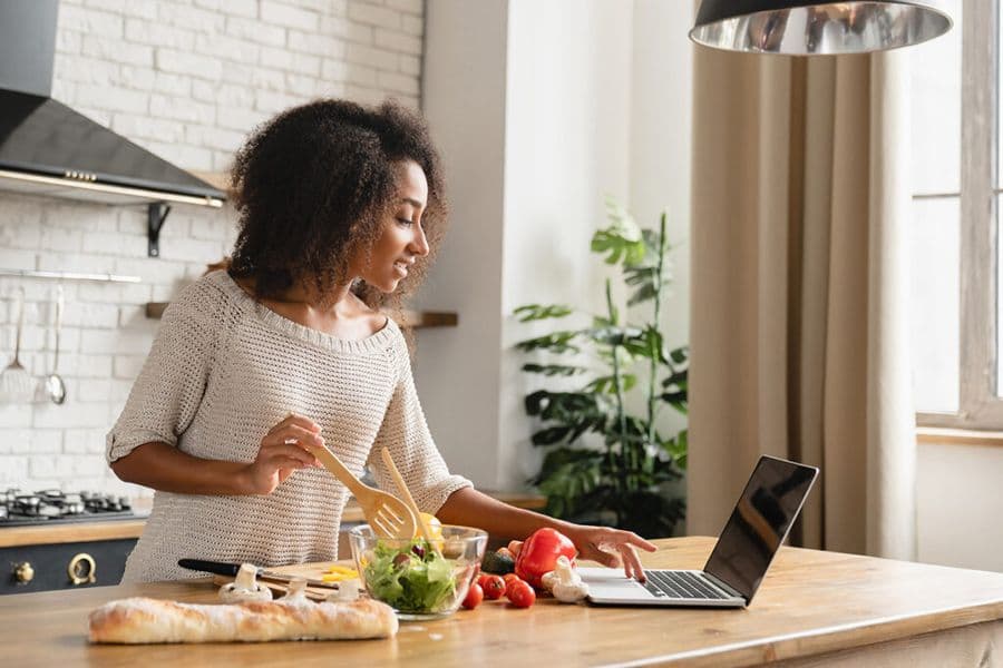 A woman is preparing a healthy meal in a modern kitchen while following instructions on a laptop.
