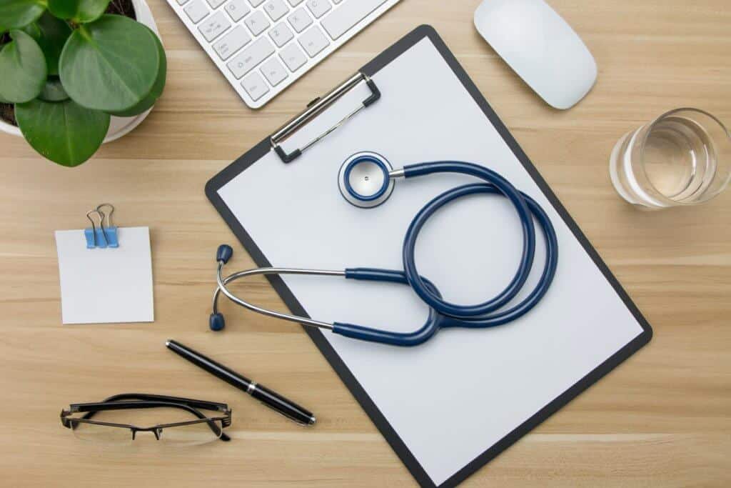 office table with stethoscope, notepad, glass of water, plant, keyboard, computer mouse, pen, and glasses