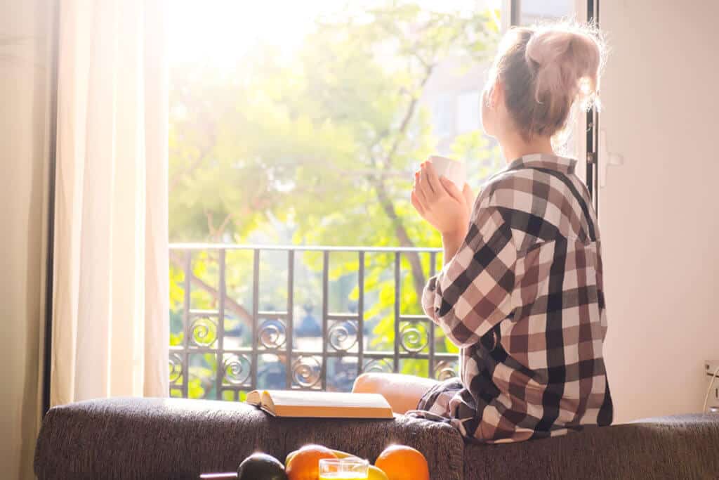 woman sitting on couch and looking out of her window as the sunny day with a mug in her hands