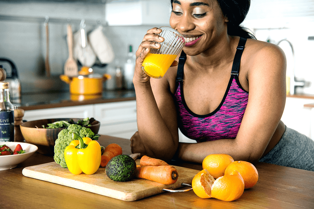 woman leaning on kitchen table drinking orange juice with raw fresh fruits and vegetables around her