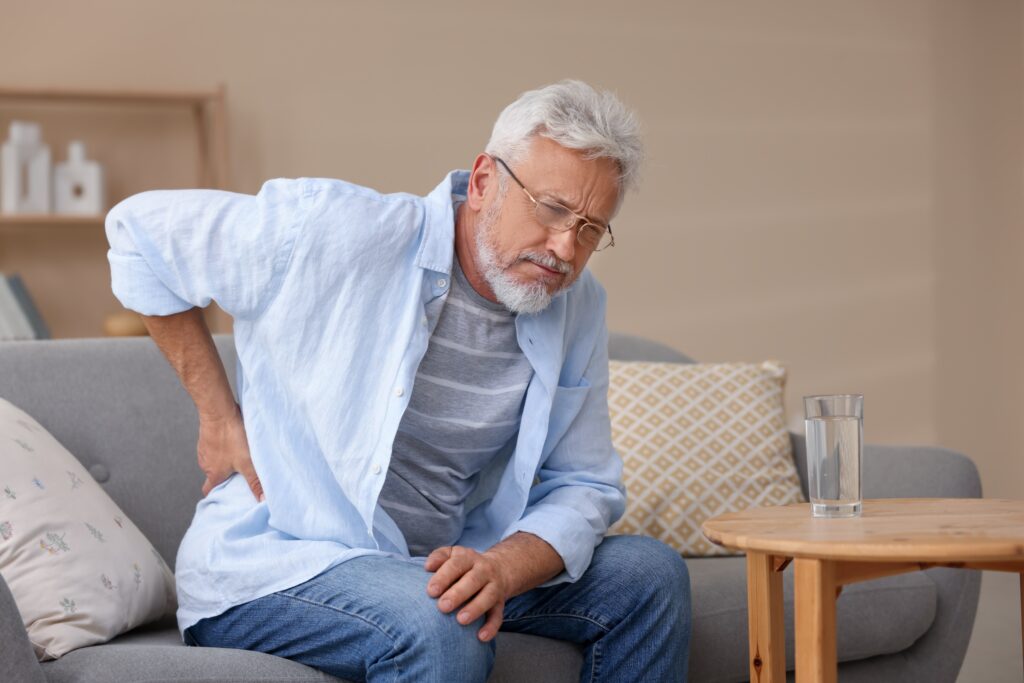 An older man is seated on a couch, holding his back in apparent pain.
