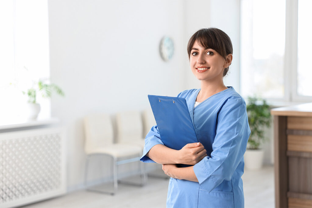 Smiling female medical assistant in blue scrubs holding a blue clipboard in a bright clinic reception area.