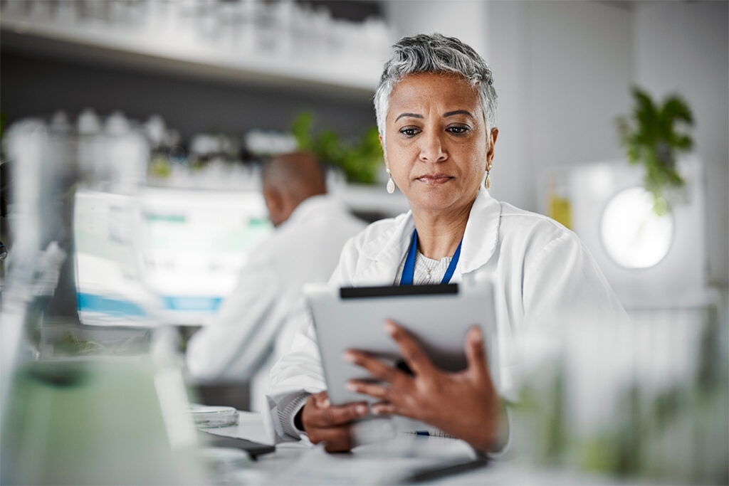 A woman in a lab coat is holding a tablet, likely engaged in her work within a laboratory setting.