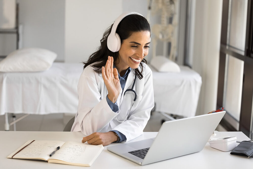 A woman is wearing headphones and waving at her laptop.