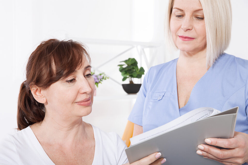 A woman is holding a book and looking at another woman. They are indoors, and both appear to be smiling.