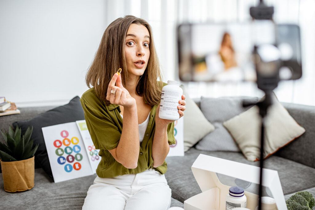 A woman is sitting indoors, holding a pill and a bottle.