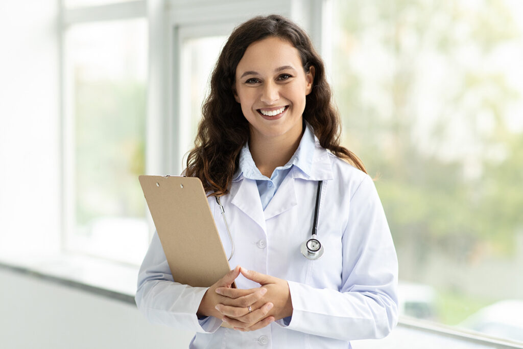 A woman is seen wearing a white coat, smiling indoors.